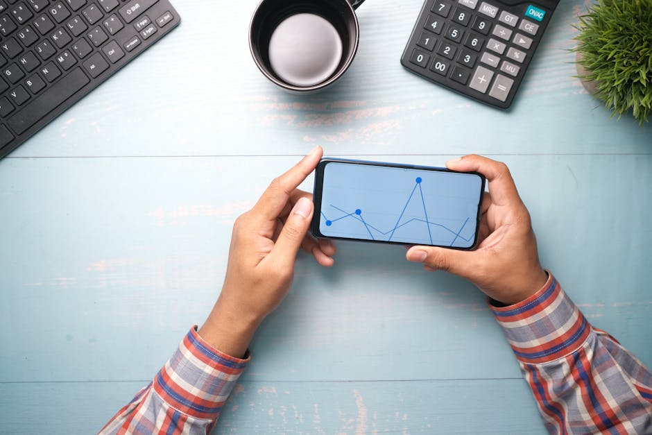 High angle view of hands holding a smartphone with a graph, surrounded by office items on a wooden table.