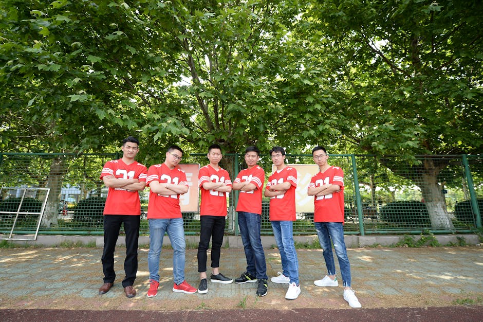 Six young men in matching red jerseys pose confidently outdoors under green trees.